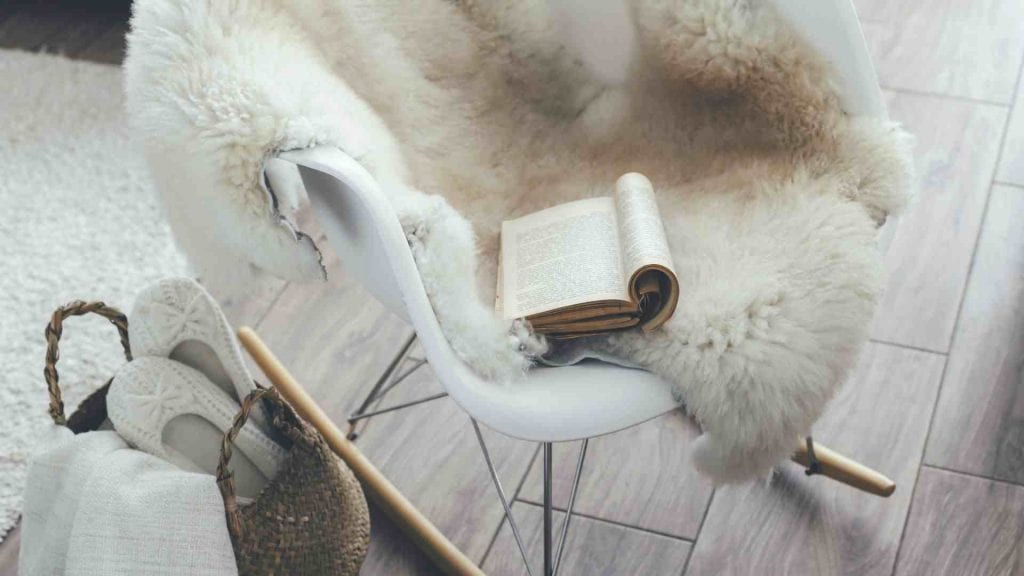 A white arm chair with a cream, fluffy rug draped over the top and an open book placed on top. The chair sits on a wooden floor with a cream, fluffy hygge rug in the background. There is a wicker basket next to the chair with comfortable white slippers and a white blanket inside. These are great examples of hygge decorating tips.